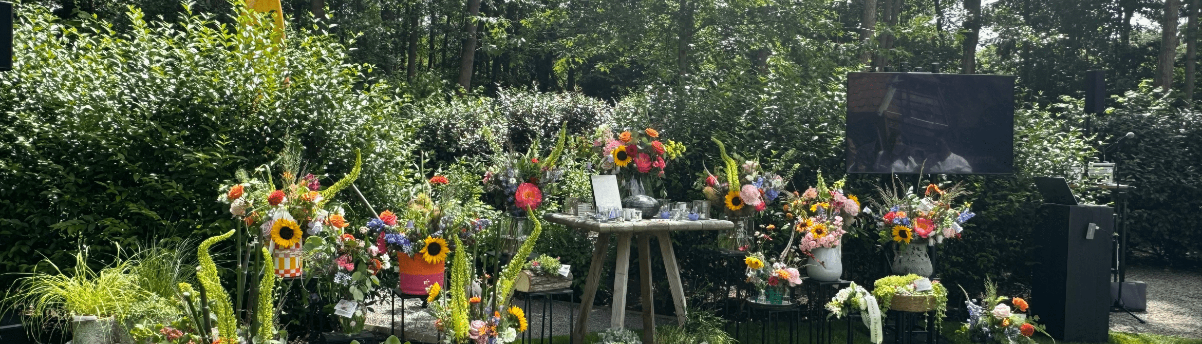 Persoonlijk ingerichte afscheidsplek in een groene tuin. Op het gras staan tientallen kleurrijke bloemstukken rond een centrale herdenkingstafel. Rechts zijn een scherm en geluidsinstallatie opgesteld voor een uitvaartceremonie in openlucht. Op de voorgrond staan witte stoelen voor de aanwezigen. Op de achtergrond vormen bomen en struiken een rustige, natuurlijke omgeving.